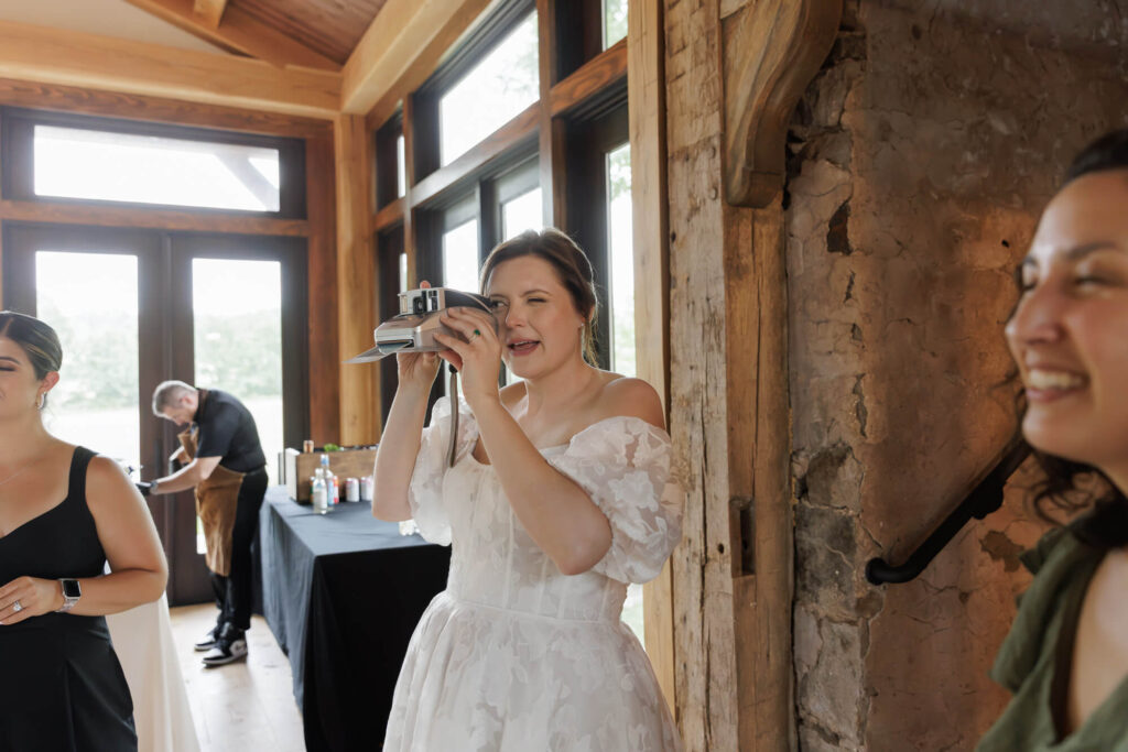 A bride in a long white gown takes a photo of friends with a Polaroid instant camera during a micro wedding reception in Northridge Barn