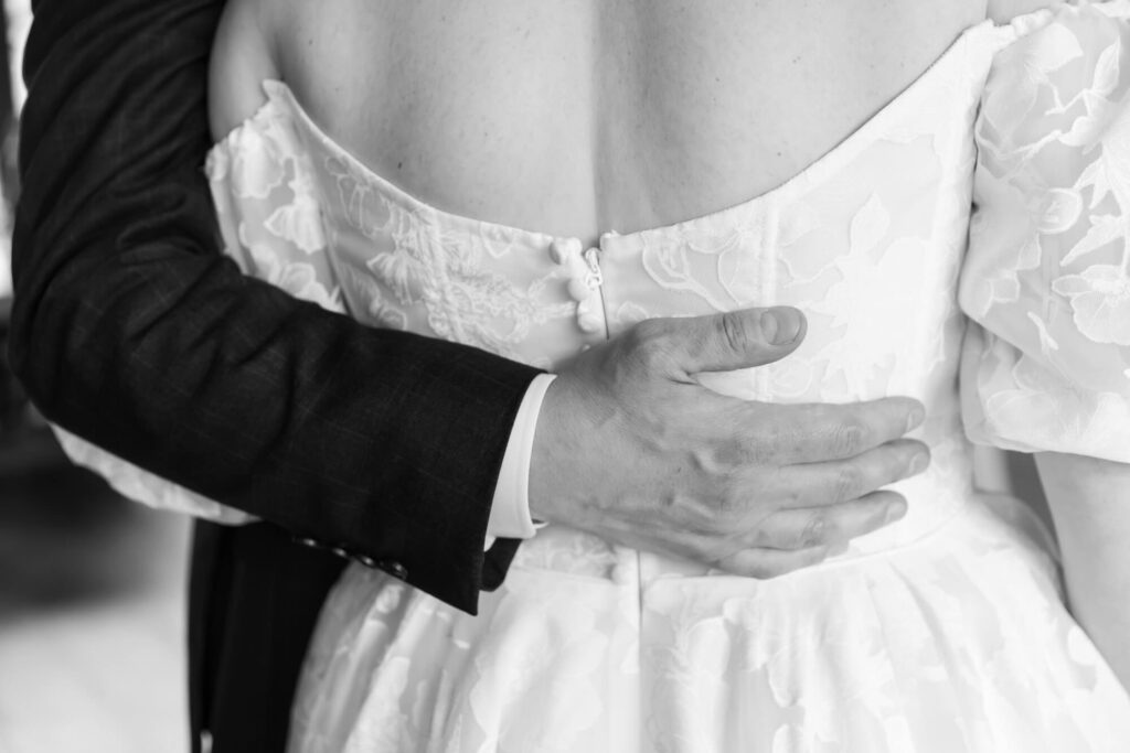 A black and white photo from behind of a groom with his arm around the waist of the bride during cocktail hour