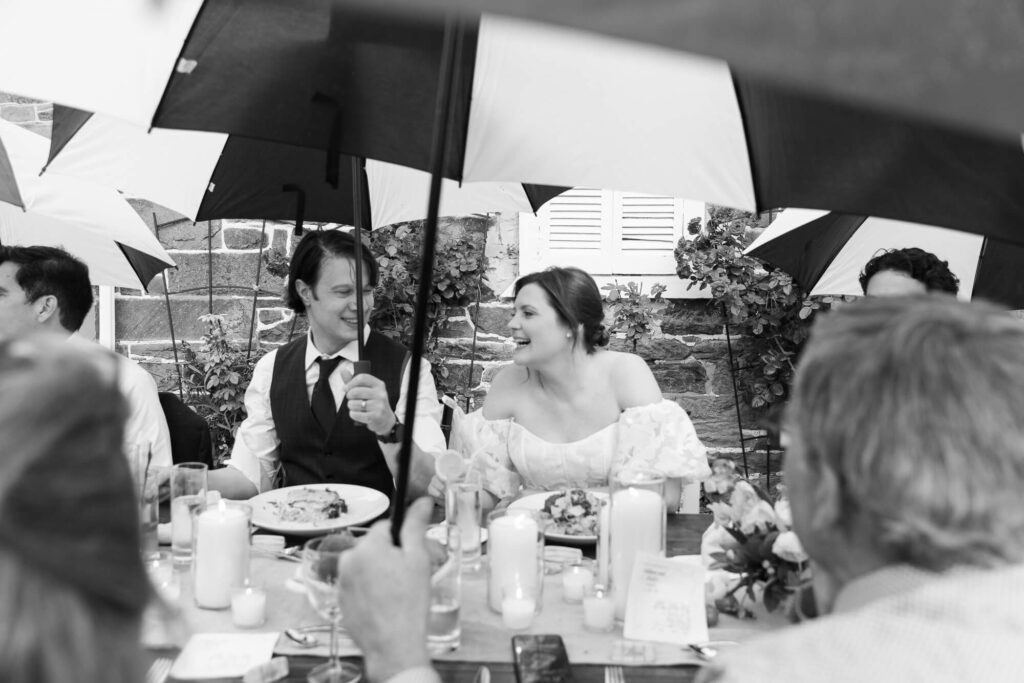 Black and white photo of a bride laughing with a groom while they are seated at their outdoor wedding dinner, holding a black and white golf umbrella to protect them from rain