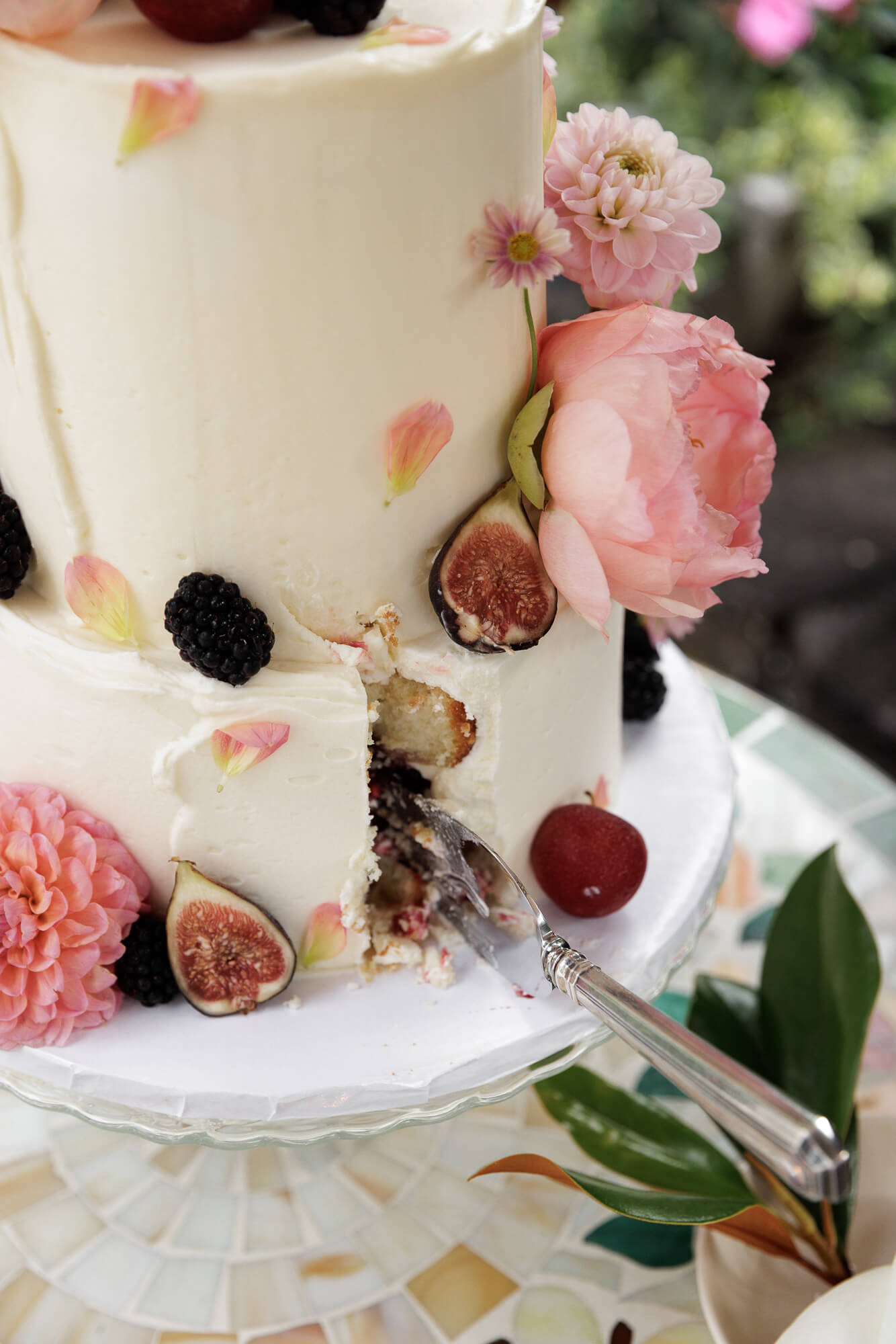 A three tier white cake decorated with pink petals, pink floral blossoms, figs and blackberries has one slice taken out, the long handled knife still inserted into the bottom layer at the missing piece