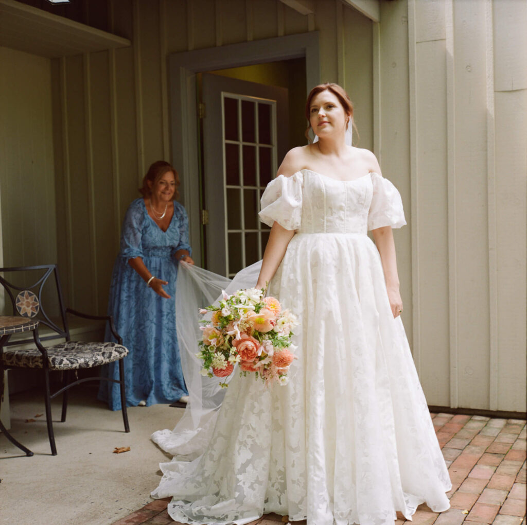 Film photograph of a bride in a full white, puffy sleeve off the shoulder gown, her mother is in the background in a light blue gown adjusting the train of the dress