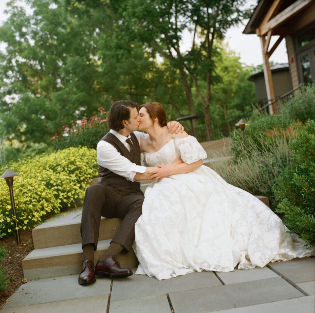 Film photograph of Bride and groom embrace each other and kiss while seated on the bluestone steps of Northridge Barn, in an outdoor landscape with yellow flowers and green leaves around them