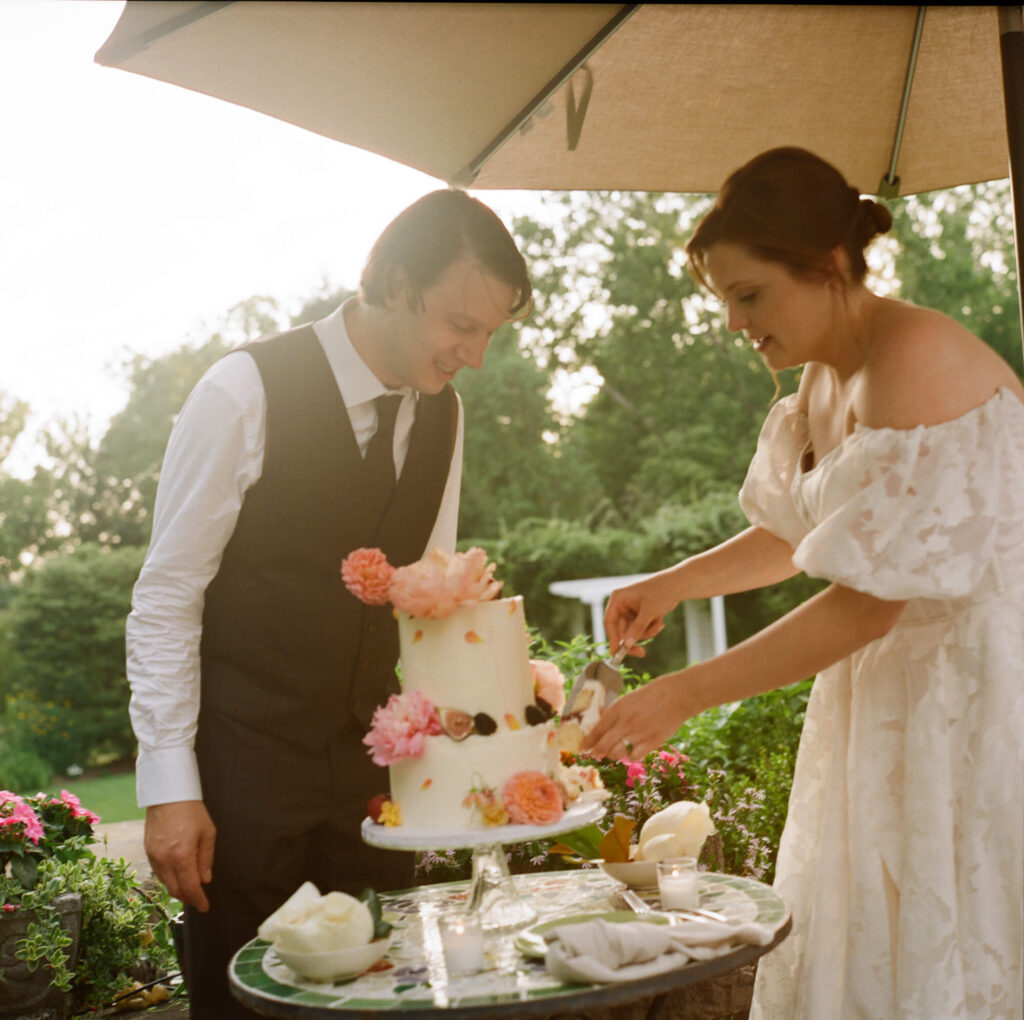 Film photo of a Groom in a white shirt, tie and formal suit vest looks on as a bride in a full, off the shoulder gown cuts a piece from a two tiered wedding cake