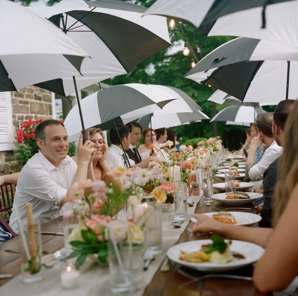 film photo of guests seated at an outdoor dinner table during a micro wedding, holding black and white golf umbrellas as they eat dinner