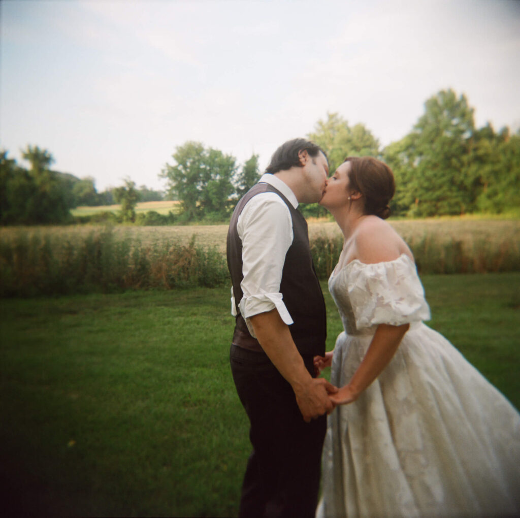 Film photo of a bride and groom standing in an open field and kissing with the treeline and a field of tall grasses behind them