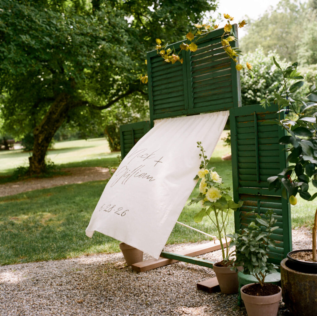 A film photo of a large, standing frame made of upcycled green shutters with a linen banner inside that says "Nick + Gillian 6.28.26"