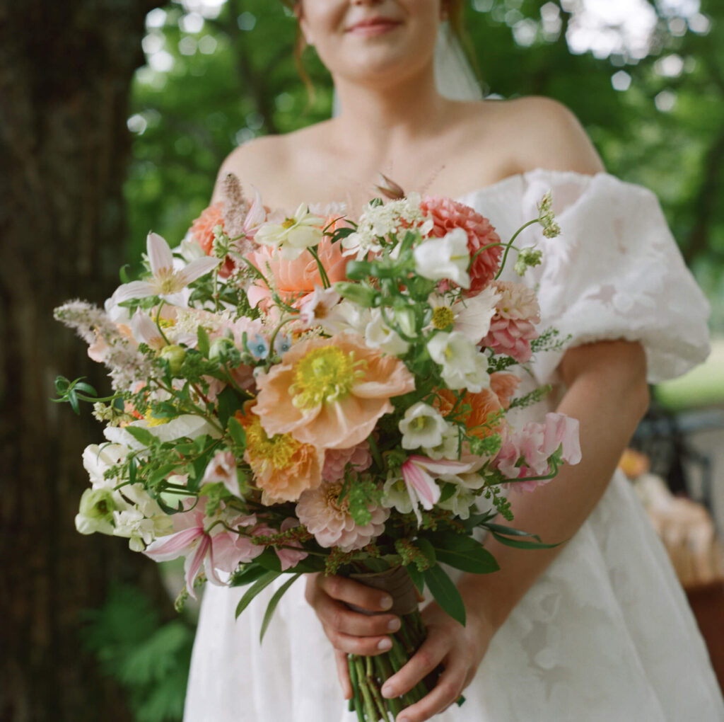 Film photograph of a large, lush bouquet of white, coral, and pink flowers being held by a bride in a puffy sleeve, off the shoulder gown