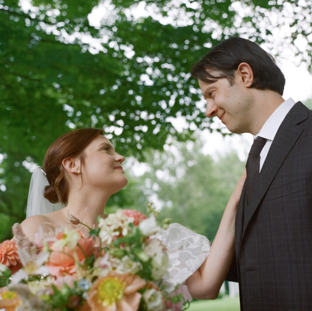 A film photograph of a bride and groom standing under tall shady trees, the bride reaches out to touch the groom on the shoulder as they look into each other's eyes