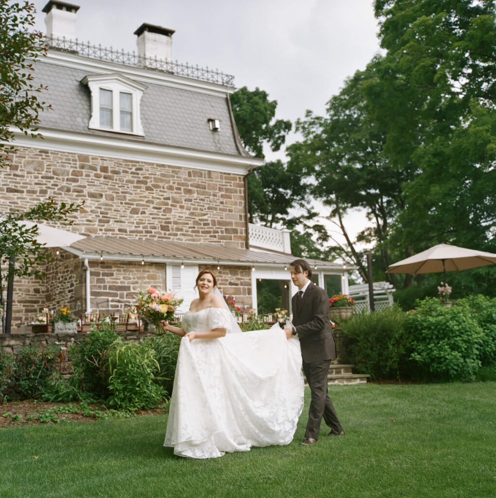 Film photograph of a bride walking on the side of the Woolverton Inn while the groom lifts the train of her dress